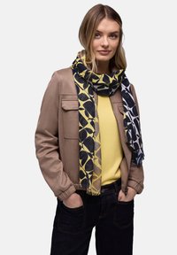 Femme portant une veste beige, un haut jaune, une écharpe à motifs bleus et un jean foncé, les mains dans les poches, sur un fond uni.