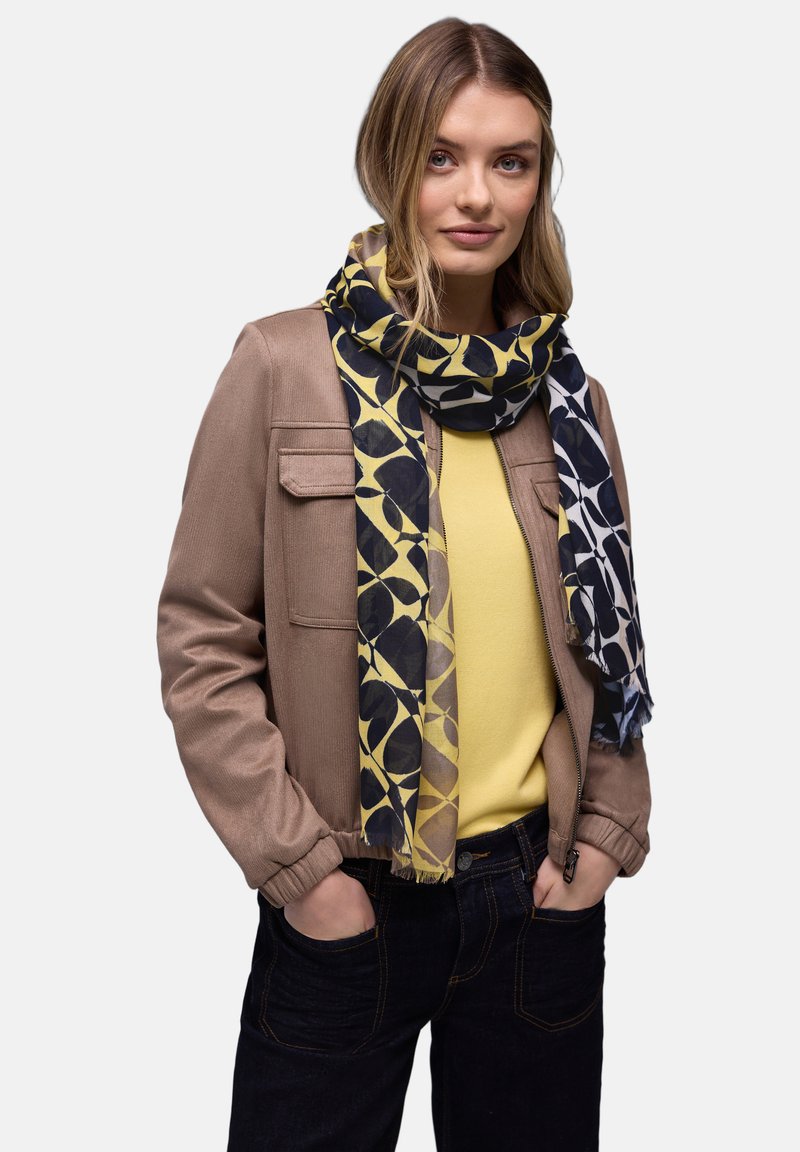 Femme portant une veste beige, un haut jaune, une écharpe à motifs bleus et un jean foncé, les mains dans les poches, sur un fond uni.