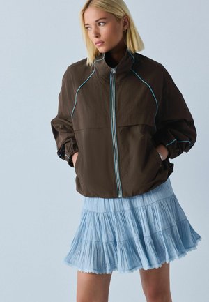 Blonde Frau trägt eine braune Jacke mit Reißverschluss und blauer Paspel sowie einen hellblauen gestuften Rock, Hände in den Taschen, schaut seitlich.