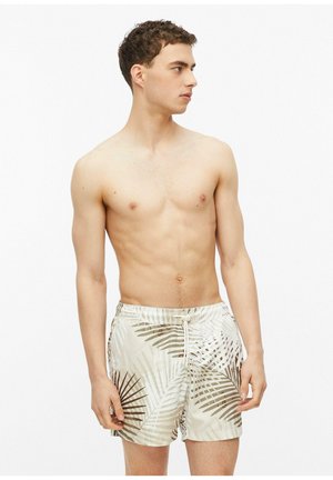 Jeune homme portant un short de bain beige avec des motifs de feuilles de palmier marron et blanc, debout sur un fond blanc uni.
