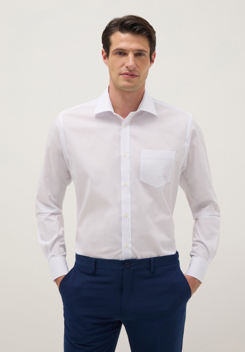 Hombre vistiendo una camisa blanca de manga larga ajustada con bolsillo frontal y pantalones azul marino, con las manos en los bolsillos, fondo neutro.