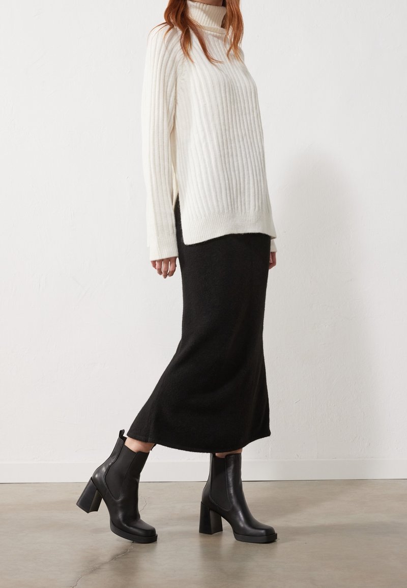 Femme portant un pull à col roulé blanc côtelé, une jupe midi noire et des bottines à talons noires, debout contre un mur blanc.