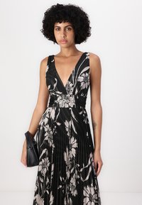 Robe noire à fleurs avec un décolleté plongeant, un design plissé et des motifs floraux clairs contrastants. Portée avec un petit sac à main noir.