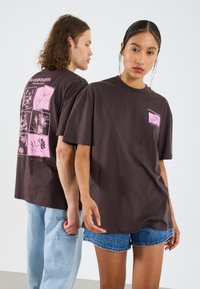 Brun bomull T-skjorte med grafiske trykk i rosa, med blomstermønster på baksiden og en liten logo foran. Korte ermer og avslappet passform.