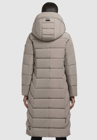 Osoba v dlhom svetlo béžovom prešívanom kabáte s veľkou kapucňou, stojaca chrbtom k nám na neutrálnom svetlom pozadí.