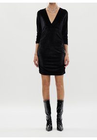 Femme portant une robe en velours noire à col en V avec des manches trois-quarts, un collier argenté remarquable et des bottes noires brillantes montant jusqu'aux genoux.