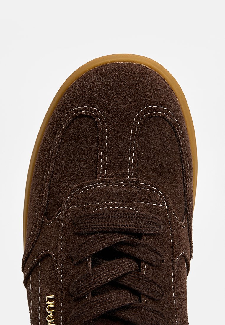 Brązowe zamszowe sneakersy z okrągłym noskiem, białymi przeszyciami i gumową podeszwą. Sznurówki są przewleczone przez wyściełany obszar języka.