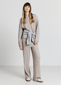 Beiger Strickpullover mit weitem Ausschnitt, kombiniert mit passenden locker sitzenden Hosen. Ein hellgrauer Sweatshirt ist um die Taille gebunden.