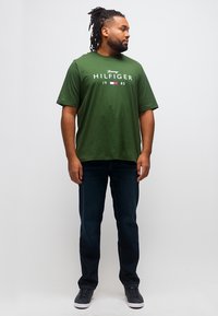 Άνδρας με dreadlocks που φοράει πράσινη μπλούζα Tommy Hilfiger, σκουρόχρωμο τζιν και μαύρα αθλητικά παπούτσια, στέκεται και κοιτάζει πλάγια.