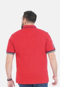 Polo rouge avec col, présentant des accents bleu marine sur les manches et le col. Texture douce, design uni sans motifs visibles.