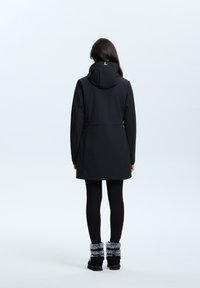 Veste noire imperméable avec capuche, présentant un design épuré et une coupe mi-longue. Associée à des leggings noirs et des bottes avec accents en fourrure.