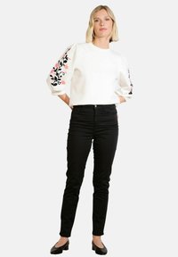 Donna che indossa una blusa bianca con ricami floreali sulle maniche a sbuffo, pantaloni skinny neri e scarpe nere con tacco basso, in piedi con le mani dietro la schiena.