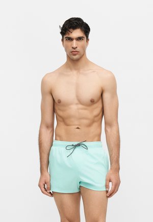 Muscular man standing facing forward wearing light blue swim shorts with black and white drawstring on plain white background.