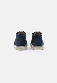 Stivaletti alla caviglia in suede blu navy con dettagli perforati, suole in gomma bianca e rivestimento interno beige. Vista posteriore che mostra un design elegante.