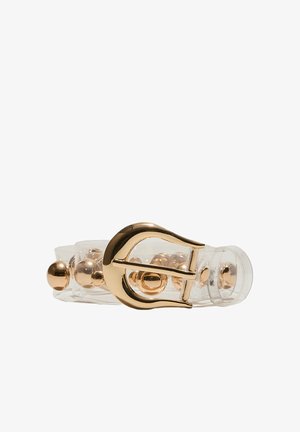 Cintura trasparente con fibbia dorata e borchie sferiche dorate distribuite uniformemente all’interno della cinghia, posata distesa su sfondo bianco.