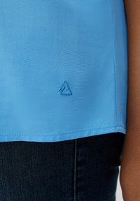 Light blue top with a smooth texture and a rounded hem. Features a small triangle logo embroidered in a darker blue. Styled with dark denim.