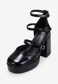 Tacchi a piattaforma neri in pelle sintetica con punta arrotondata, cinturini alla caviglia e un tacco alto e chunky. Presentano dettagli in fibbia metallica.