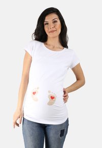 Weißes Maternity-T-Shirt mit kurzen Ärmeln, das bedruckte Babyfußabdrücke in Beige mit roten Herzen zeigt, getragen über blauen Jeans.