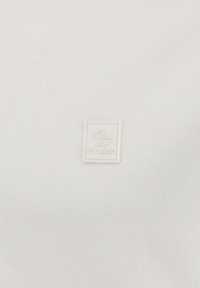 Etiqueta cuadrada blanca pequeña con un logo circular y el texto "KEY LARGO" sobre un fondo de tela blanca texturizada.