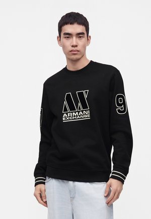 Νέος άνδρας που φοράει μαύρο φούτερ Armani Exchange με λευκά λογότυπα και τον αριθμό 9 στο μανίκι, συνδυασμένο με ανοιχτό μπλε τζιν.