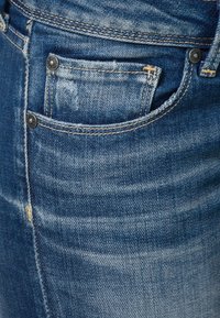 Jeans de mezclilla azul con una textura deslavada, que cuentan con un bolsillo delantero con remaches de metal y acentos de costura amarilla.