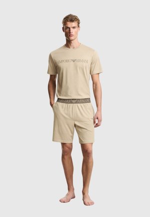 Uomo che indossa una t-shirt beige Emporio Armani e pantaloncini coordinati, in piedi a piedi nudi con le mani parzialmente nelle tasche su uno sfondo grigio chiaro.