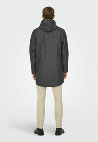 Jachetă gri impermeabilă cu glugă, având o croială dreaptă și mâneci lungi, asortată cu pantaloni subțiri de culoare deschisă și pantofi negri.