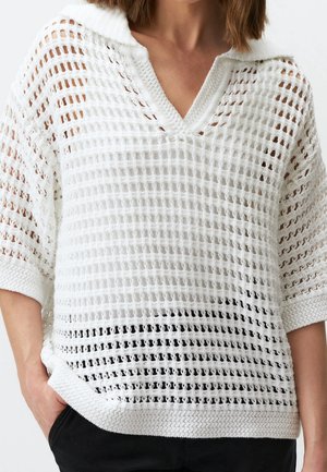 Femme portant un pull blanc à manches courtes en maille lâche avec un col en V, associé à un pantalon noir, main dans la poche.
