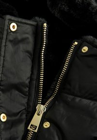 Veste en duvet noire avec une finition brillante, dotée de quincaillerie dorée, d'une fermeture éclair et de boutons-pression. Intérieur doublé de doux fausse fourrure noire.