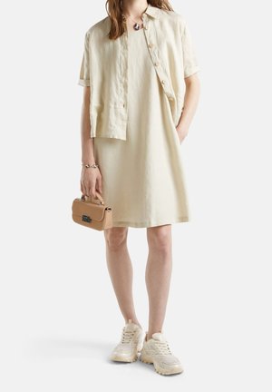 Nainen, jolla on beige lyhythihainen mekko ja siihen sopiva lyhyt napitettava paita, valkoiset paksut lenkkarit ja pieni beige käsilaukku kädessä.