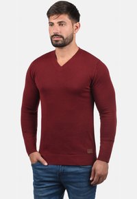 Burgunderfarbener V-Ausschnitt-Pullover aus weichem Material mit gerippten Bündchen und Saum, versehen mit einem kleinen Markenpatch unten links.