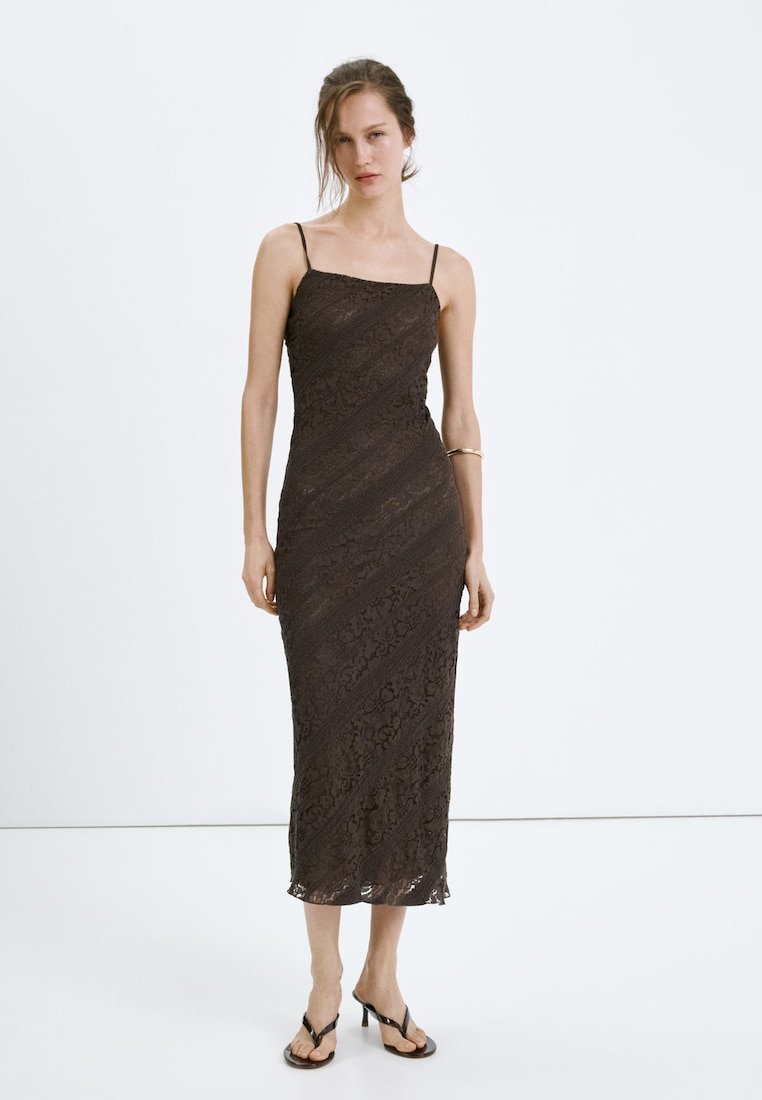 Femme portant une robe midi en dentelle marron foncé ajustée avec de fines bretelles et des sandales à talons noires, debout devant un fond blanc uni.