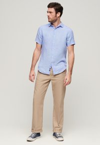 Camisa de lino de manga corta en azul claro con cuello abotonado, combinada con chinos color beige y zapatillas oscuras azules, de pie frente a un fondo neutro.