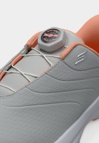 Chaussures Skechers avec tige texturée grise, accents orange et système de fermeture twist-fit. Dotées de lacets blancs et d'un logo distinctif.