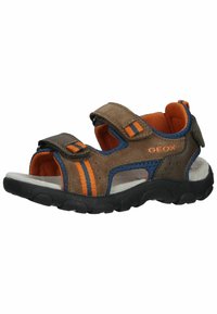 Sandalia marrón y naranja con correas de Velcro, acentos de malla y una plantilla acolchada. Cuenta con una suela negra de goma resistente y recortes laterales.