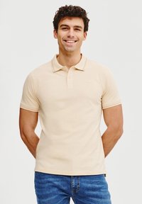 Beżowa koszulka polo z krótkim rękawem i klasycznym kołnierzykiem, z dwiema guzikami w stójce oraz subtelnym logo na piersi, w połączeniu z niebieskimi dżinsami.