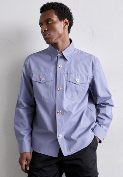 Camisa de algodón de color púrpura claro con un collar estructurado, bolsillos frontales y botones en tono dorado, que presenta mangas largas y un ajuste relajado.