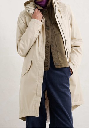 Personne portant un manteau à capuche beige par-dessus une chemise marron à boutons et un pantalon en velours côtelé bleu marine, tenant le col du manteau d’une main.