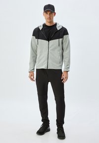 Sporty zip-up jakke i grå og sort, lavet af blødt stof, med hætte, snore og lomme foran. Sammen med sorte joggers og sneakers.