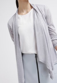 Jasnoszary kardigan o miękkim, drapowanym fasonie, nałożony na biały top, zestawiony z jasnoniebieską spódnicą z zapięciem na zamek.