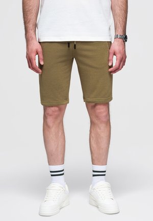 Mand stående i khakishorts, hvid T-shirt, stribede hvide sokker og hvide sneakers, med hænderne afslappet ned langs siderne, synlig til midt på låret.