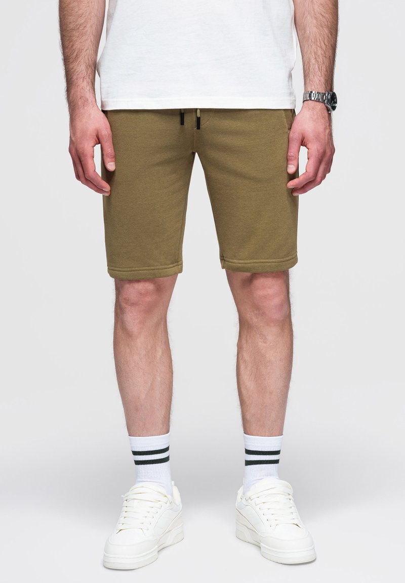 Mężczyzna stojący w szortach khaki, białej koszulce, białych prążkowanych skarpetach i białych sneakersach, z rękami swobodnie opuszczonymi wzdłuż ciała, widoczny do połowy uda.