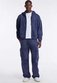 Marineblauwe hoodie met een rits aan de voorkant, gecombineerd met bijpassende cargobroek met twee zijzakken. Draagt over een wit T-shirt.