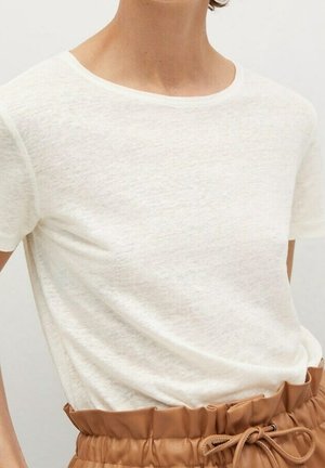 Personne portant une chemise blanche unie à manches courtes rentrée dans un pantalon beige taille haute avec une taille froncée et une ceinture nouée.