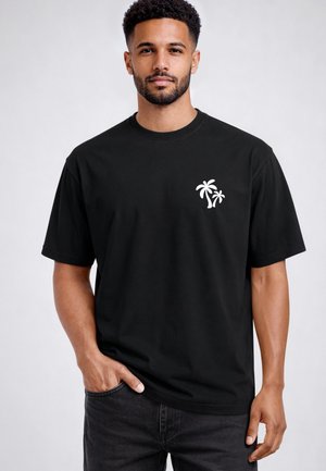 Mann trägt ein schwarzes T-Shirt mit kleinem weißem Palmen-Grafik auf der linken Brust, steht mit einer Hand in der Tasche vor grauem Hintergrund.