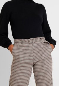 Černý rolák spárovaný s hnědými a bílými kostkovanými kalhotami s vysokým pasem, které mají detail vázaného opasku a boční kapsy.