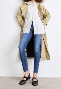 Gabardina beige con botones, camisa blanca a rayas, jeans ajustados azules y zapatos de malla negros con correa y acentos. Texturas suaves.