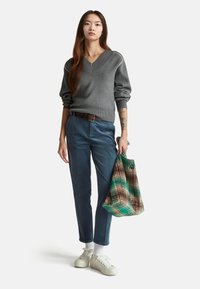 Grau gestrickter Pullover mit V-Ausschnitt, blaue Cordhosen, weiße Sneakers und eine mehrfarbige Tragetasche mit Batikmuster.