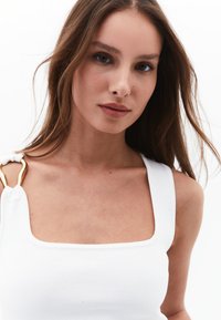 Top bianco a righe con scollatura quadrata, caratterizzato da un accento dorato sulla spalla. Texture liscia, design aderente.