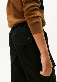 Zwarte cargobroek met zijzakken, voorzien van een logo-opdruk. Het model draagt een bruine langemouwen t-shirt. Elastische tailleband zichtbaar.